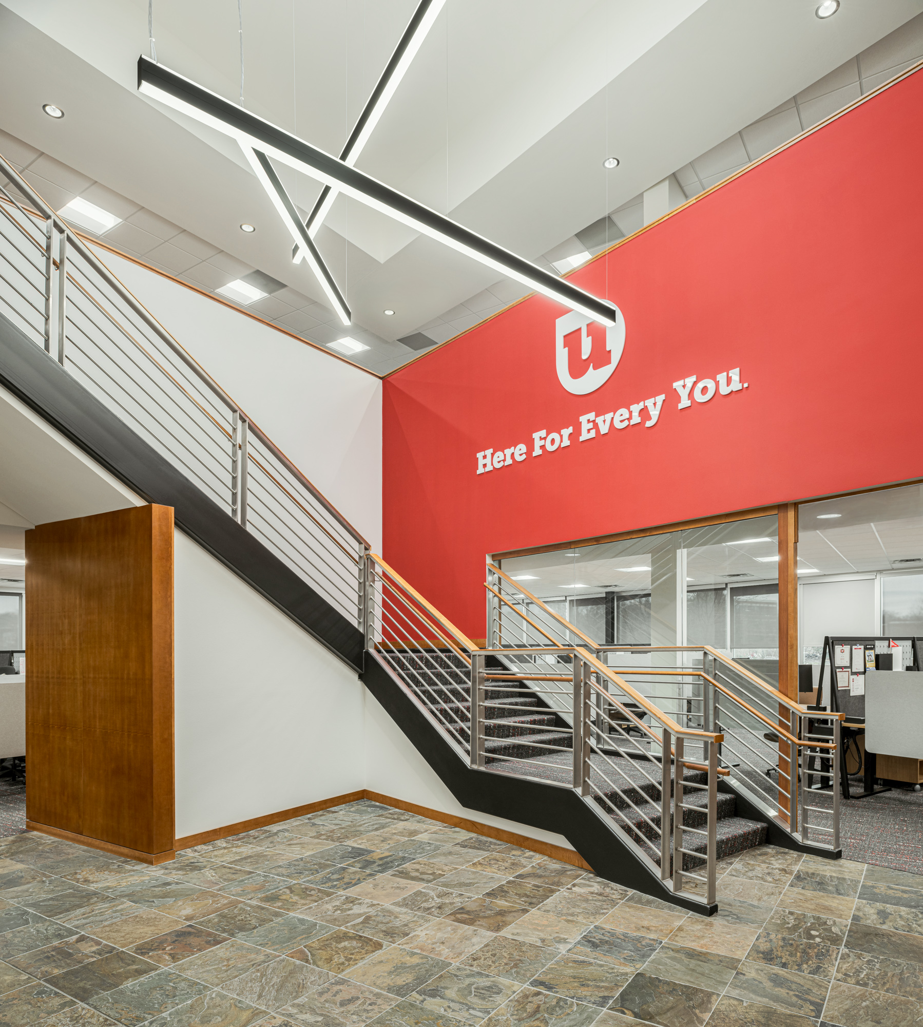 A modern office interior with a red feature wall displaying a white logo and the text "Here For Every You." The space includes a sleek, metal-framed staircase with wooden handrails. The floor is tiled, and an array of overhead lights provides illumination.