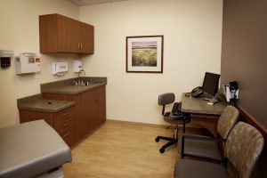 A medical examination room with a patient bed, a wall cabinet, a sink with countertops, and a desk with a computer, phone, and chair. There are two additional chairs in the room and a framed picture hanging on the wall above the desk.