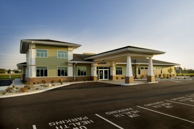 A single-story healthcare facility with a wide entrance and ample parking spaces labeled for health services in front. The modern building features large windows, a mix of green and beige exteriors, and landscaped surroundings with shrubs and white stones.