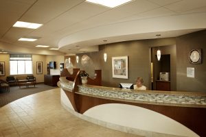 A modern, spacious office reception area with a curved desk. A receptionist is seated behind the desk, using a computer. The decor includes neutral tones, hanging lights, a large clock, and artwork on the walls. To the left are chairs and a few tables in a waiting area.