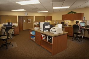 A spacious office with a central island holding printers and office supplies. Two people work at their desks along the walls. The room is well-lit with fluorescent lights, and there is a clock on the wall. Various office supplies and documents are neatly organized.