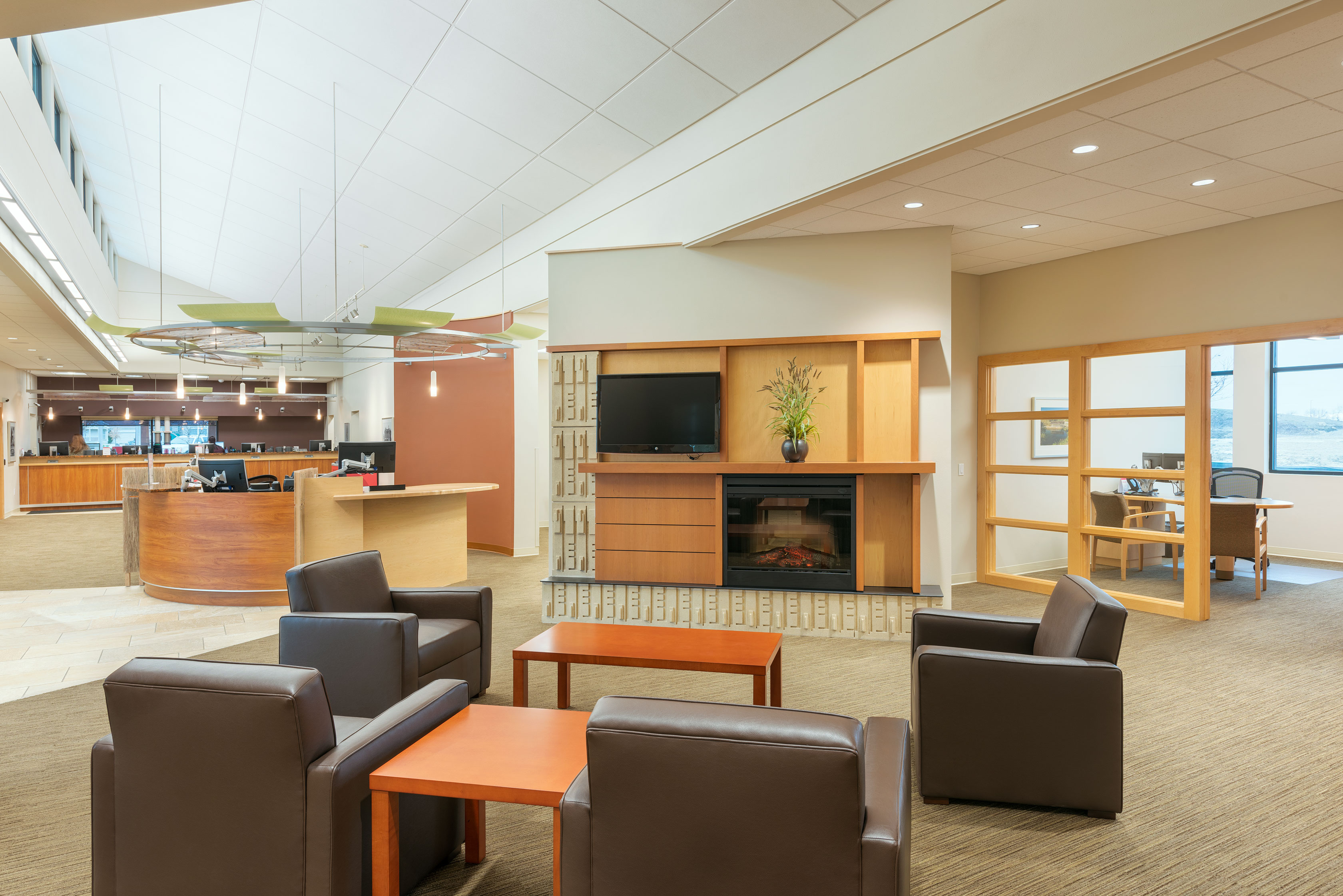 A modern waiting area with four dark leather chairs arranged around two wooden tables. A built-in fireplace and a TV are set into a wooden feature wall. The space is open with a reception desk in the background and an office visible through glass doors on the right.