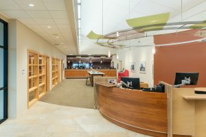 modern office lobby with a curved wooden reception desk equipped with two computer monitors. Shelves line the wall to the left. The carpeted floor leads to a seating area with red and beige chairs. Pendant lights and framed art adorn the space.