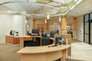 A modern, well-lit office reception area featuring a large wooden desk with computer stations. Behind the desk are cabinets and shelves with decor. Ceiling lights and stylish green and beige panels hang overhead. To the right is a glass door entrance.