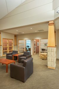 A modern office space with brown leather armchairs, a wooden coffee table, beige walls, and brown carpeting. In the background, there are glass doors leading to offices and a small desk with chairs. The area is well-lit with overhead lighting and natural light.