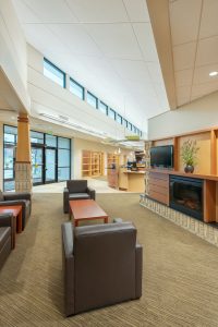 A modern, well-lit lobby area featuring several dark brown leather chairs and wooden tables. The space includes large windows, a mounted flat-screen TV, built-in bookshelves, and a fireplace. The interior design is enhanced with neutral colors and recessed lighting.