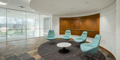 A modern office lounge area with four teal swivel chairs arranged around a small round table on a circular rug. The space has a curved wooden accent wall and large windows offering a view of trees and parked cars. The floor is tiled in a slate pattern.