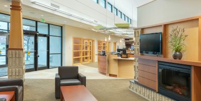 well-lit lobby area featuring several dark brown leather chairs and wooden tables. The space includes large windows, a mounted flat-screen TV, built-in bookshelves, and a fireplace. The interior design is enhanced with neutral colors and recessed lighting.