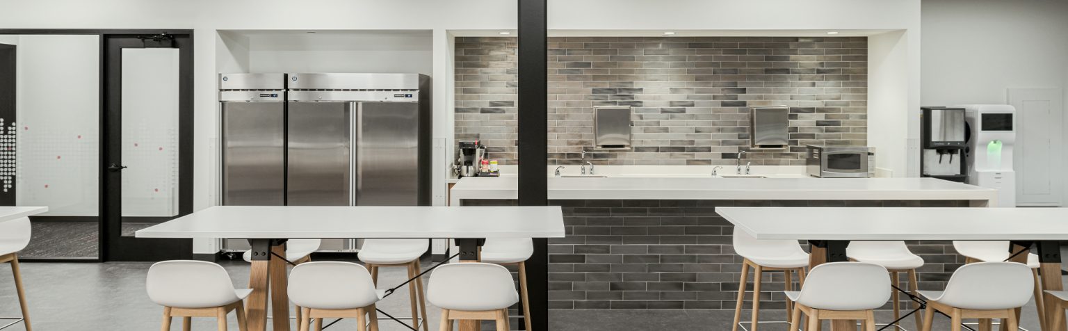 minimalist kitchen area featuring white tables and stools with light wooden legs. The space has stainless steel refrigerators on the left, a coffee machine and sinks in the back, and a backdrop of grey brick tiles. Walls and ceiling are white.