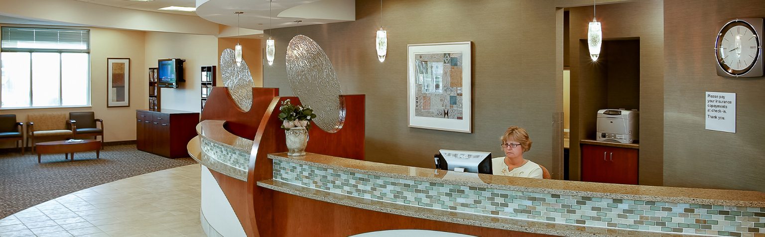 A modern, spacious office reception area with a curved desk. A receptionist is seated behind the desk, using a computer. The decor includes neutral tones, hanging lights, a large clock, and artwork on the walls. To the left are chairs and a few tables in a waiting area.