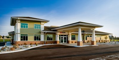 A single-story healthcare facility with a wide entrance and ample parking spaces labeled for health services in front. The modern building features large windows, a mix of green and beige exteriors, and landscaped surroundings with shrubs and white stones.