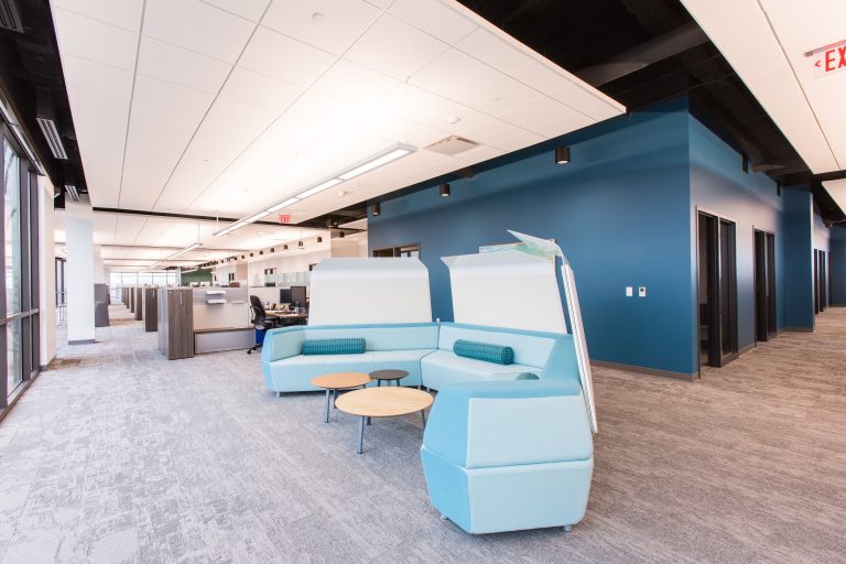 office space with blue walls and light gray carpet. In the foreground, there is a seating area with light blue modular sofas and round wooden tables. The background shows open workstations and office cubicles.