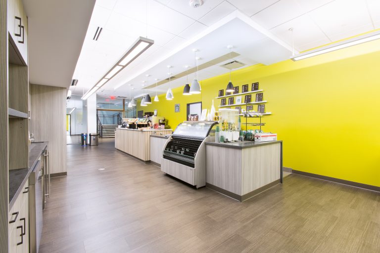 cafe interior with a vibrant yellow wall. It features a countertop with a glass display case for pastries and shelves with coffee products. Overhead pendant lights illuminate the space, and wooden flooring adds warmth.