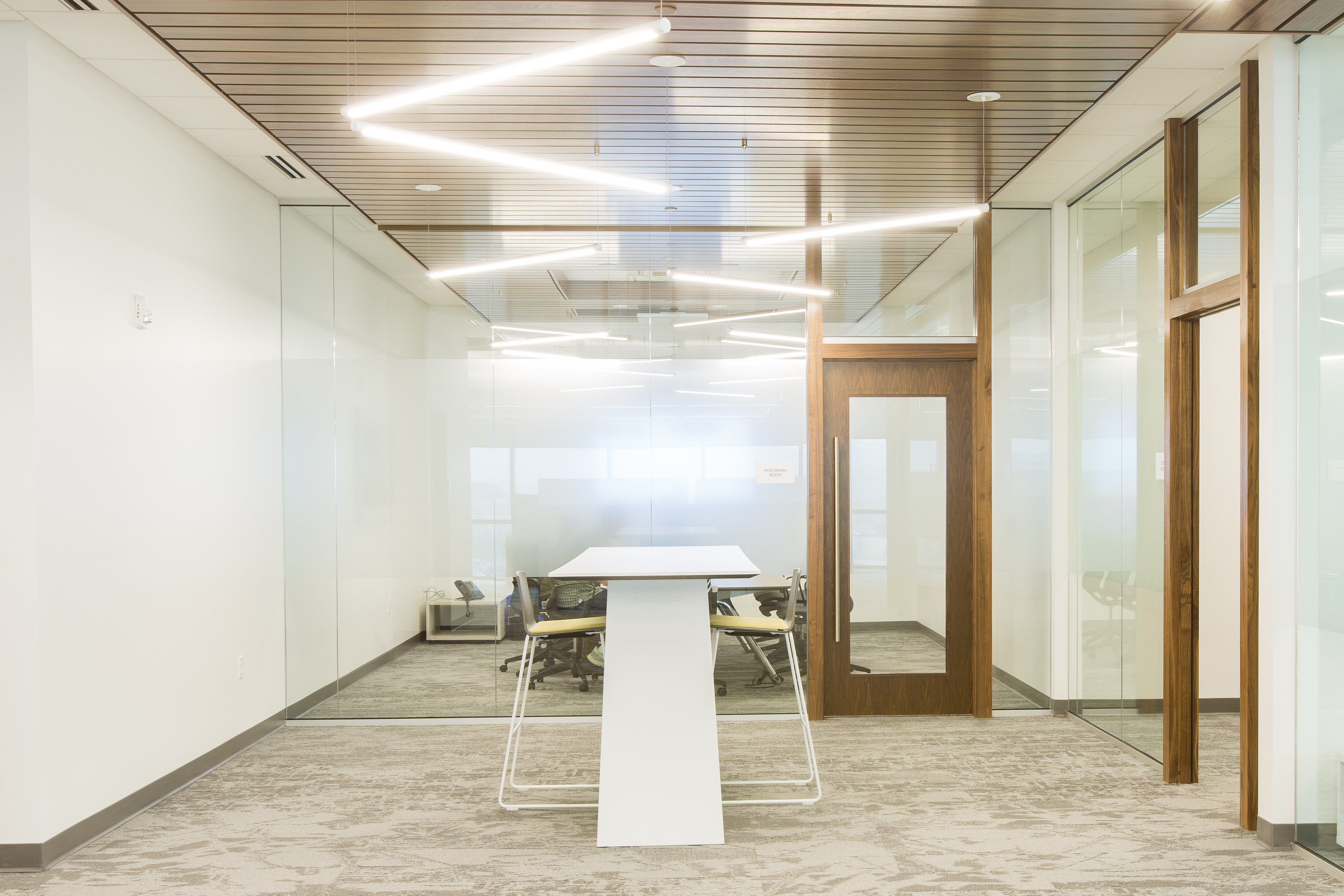 A modern office hallway with glass walls, a wooden ceiling, and zig-zagging overhead lights. A white high table stands at the center of the hallway, with a glimpse of a small meeting room containing a table and chairs visible through a glass door on the right.