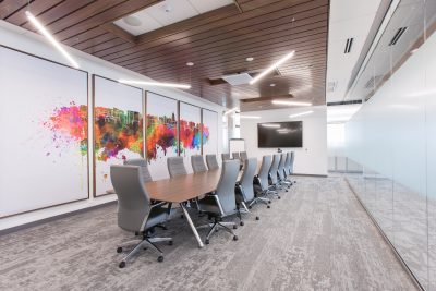 A modern conference room with a long wooden table surrounded by gray office chairs. The back wall features abstract colorful cityscape artwork.