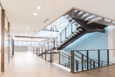 A modern office interior featuring a sleek, black steel staircase with cable railings and glass walls. The space has wooden flooring and is well-lit with recessed ceiling lights, offering a spacious, minimalist aesthetic.