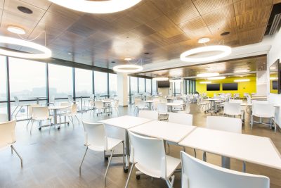 spacious cafeteria with white tables and chairs. Large windows provide natural light, and circular ceiling lights add a contemporary touch. The back wall is painted bright yellow, and multiple screens are mounted on it.