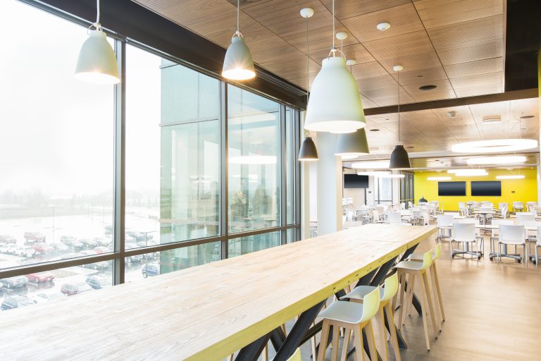 Modern office cafeteria with natural lighting, featuring a long wooden table, white chairs, and pendant lights.