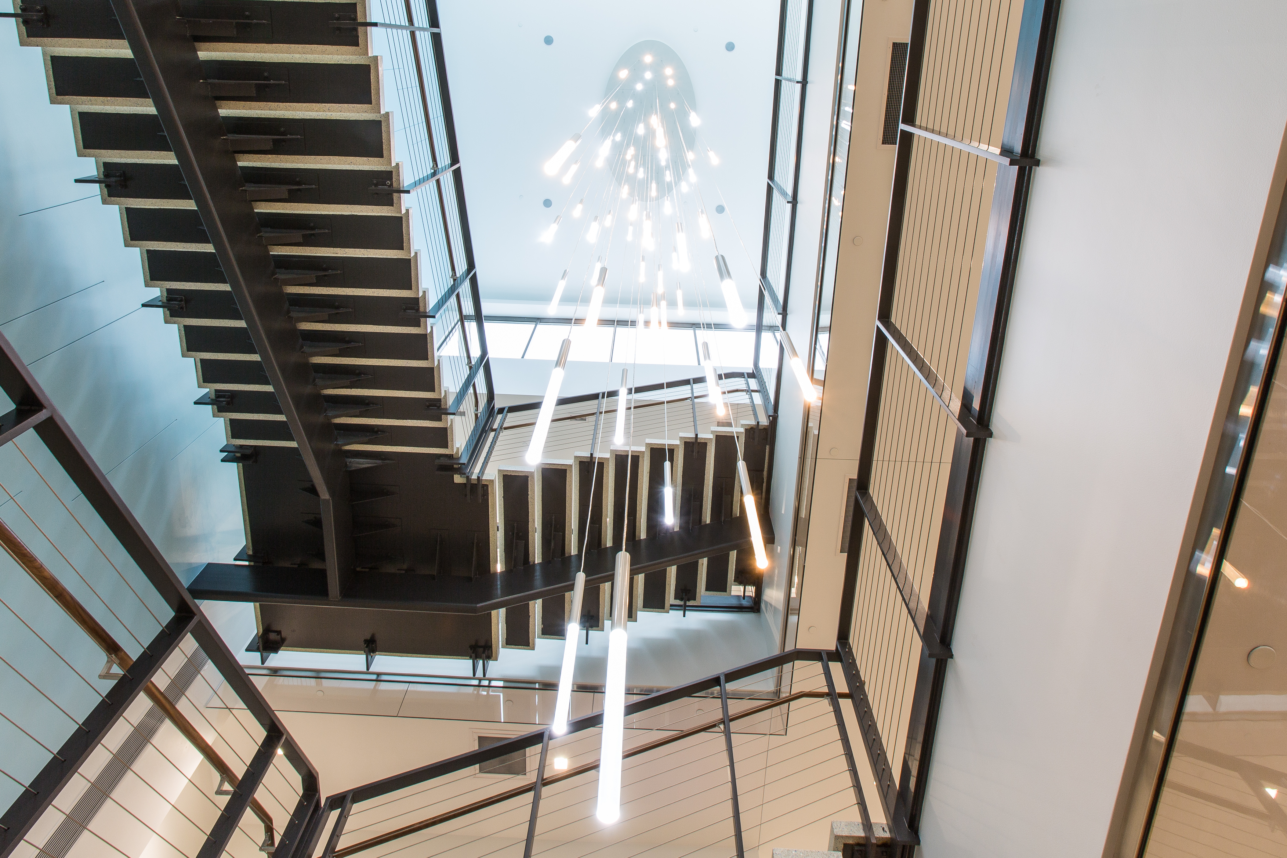 A spacious, modern lobby with white walls and large windows letting in natural light. The area features a staircase with black railings, a minimalist pendant light fixture, and glass-walled offices. The floor has light wood and tiled sections.