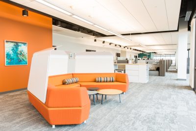 office space with an orange couch featuring striped pillows and two small tables in front. There are white dividers behind the couch. The room has an orange accent wall with a painting and open workstations in the background.