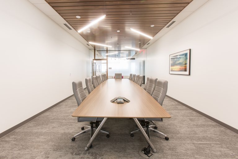 A modern conference room with a long, wooden table surrounded by gray office chairs. The ceiling has wooden paneling and modern lighting.