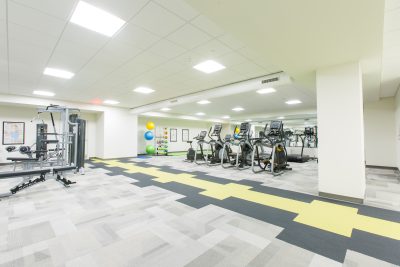 A bright, spacious gym with modern equipment, including treadmills, exercise bikes, and a multi-functional weight machine. The room has a patterned carpet and a colorful stack of exercise balls in the corner. Large mirrors line the walls.