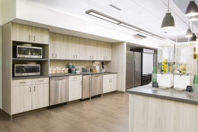 A modern kitchen with light wood cabinets, stainless steel appliances including microwaves, a dishwasher, and a refrigerator. The countertop holds glass containers filled with fruit and water, and pendant lights hang from the ceiling. The floor is a light wood.