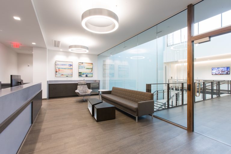 office lobby with a gray sofa, armchair, and coffee tables. The space has wood flooring, art on the walls, and glass partitions. Circular ceiling lights illuminate the area. Stairs and a hallway are visible through the glass.