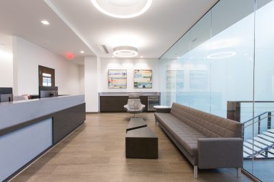 office waiting area with a gray couch, armchair, and geometric coffee table. Glass walls to the right, reception desk to the left.