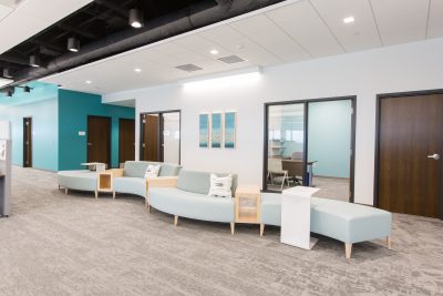 A modern office lobby with a long, curved light blue sofa and wooden side tables. The walls are painted white and teal, with art pieces hanging on the wall. Conference rooms with glass doors are in the background.