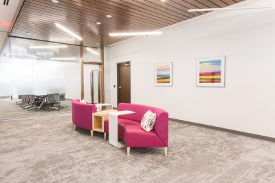 Modern office lobby with pink seating and colorful pillows, white walls adorned with abstract paintings. Glass partition leads to a conference room. Ceiling lights and carpet flooring create a bright, inviting atmosphere.