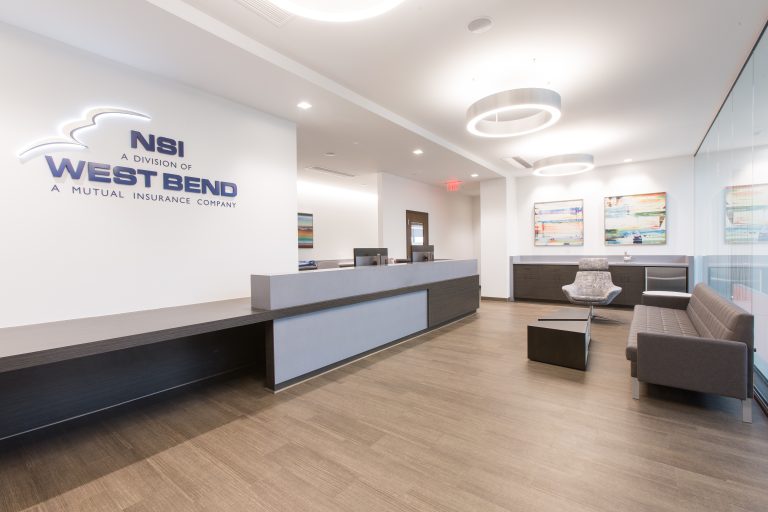 Modern office lobby with a reception desk on the left The room has a gray floor, seating area with chairs, and colorful artwork on the walls.