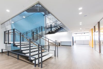 office interior featuring a sleek staircase with black railings and white steps. The stairs are enclosed by glass walls. The room has wooden flooring, a blue accent wall, and bright overhead lighting