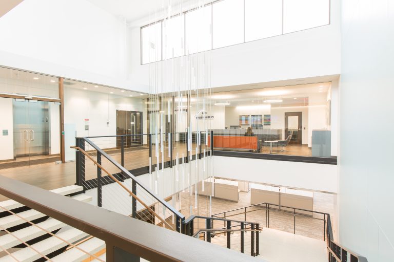 office interior with white walls and large windows, featuring a staircase with a metal railing. Offices and a lounge area with orange seating are visible through glass walls.