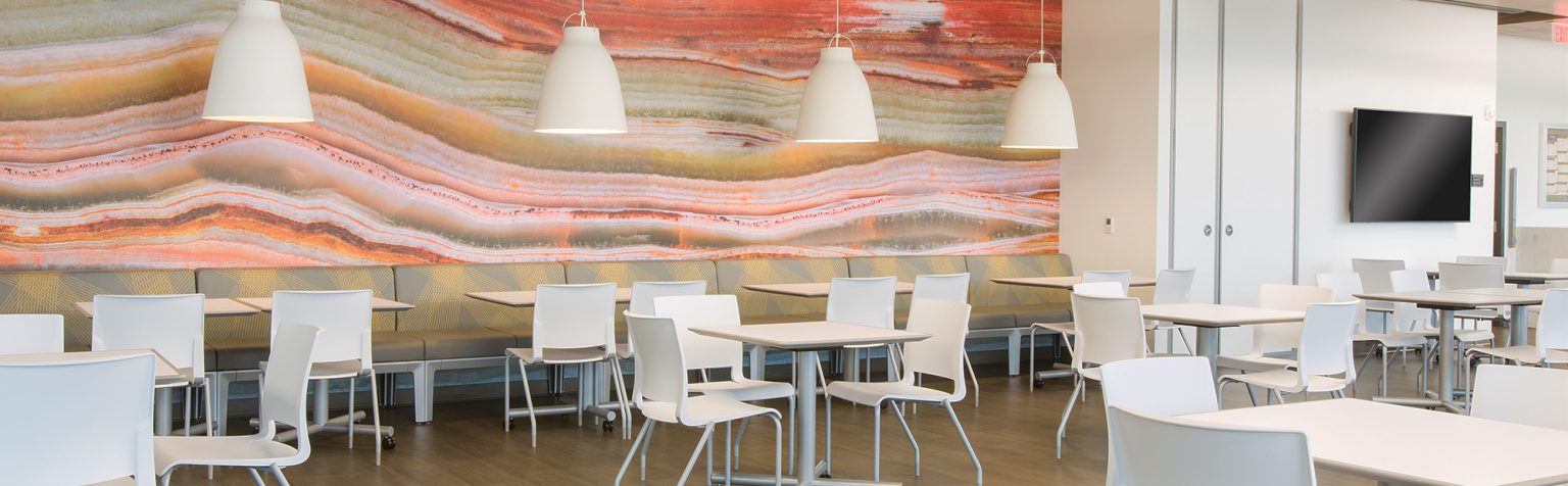 A modern cafeteria with white tables and chairs, featuring a large, colorful mural of layered earth tones on the wall. Circular ceiling lights hang above, and the floor is a light wood finish.