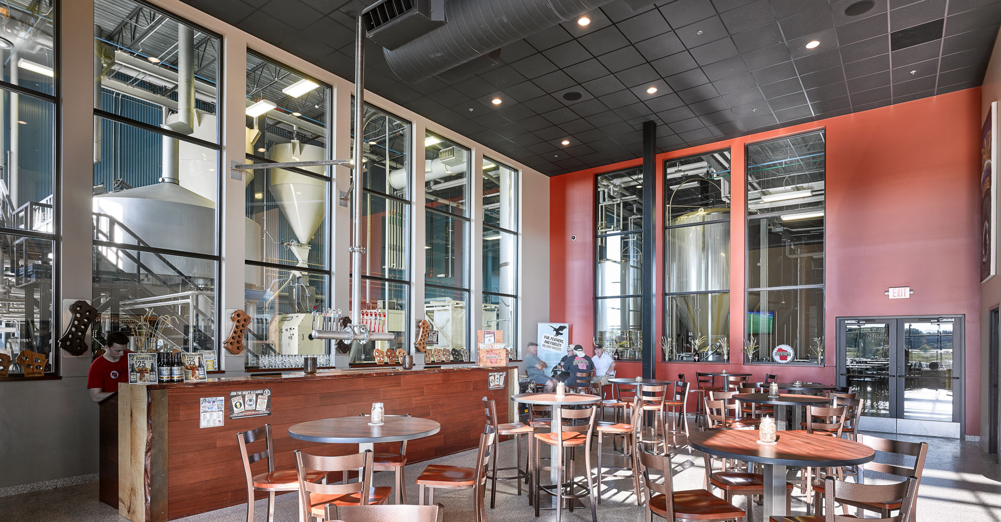 A spacious brewery taproom with large windows revealing brewing equipment. The room has high ceilings with black tiles, red and beige walls, and wooden tables and chairs arranged for seating. A wooden bar counter is adorned with various items and a chalkboard menu.