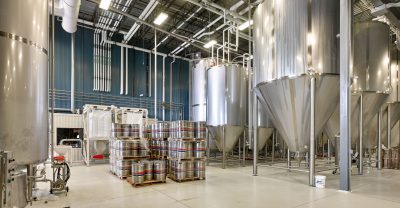 A modern brewery interior with large stainless steel fermentation tanks, stacks of metal kegs on pallets, and a clean, organized production area. The space is well-lit with overhead industrial lighting, and the floor is spotless.