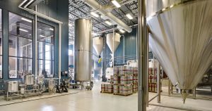 A spacious brewery with large, stainless steel fermentation tanks, kegs stacked on pallets, and various brewing equipment. The bright and clean space features high ceilings with exposed pipes and ducts, and a glass wall providing a view of adjoining rooms.