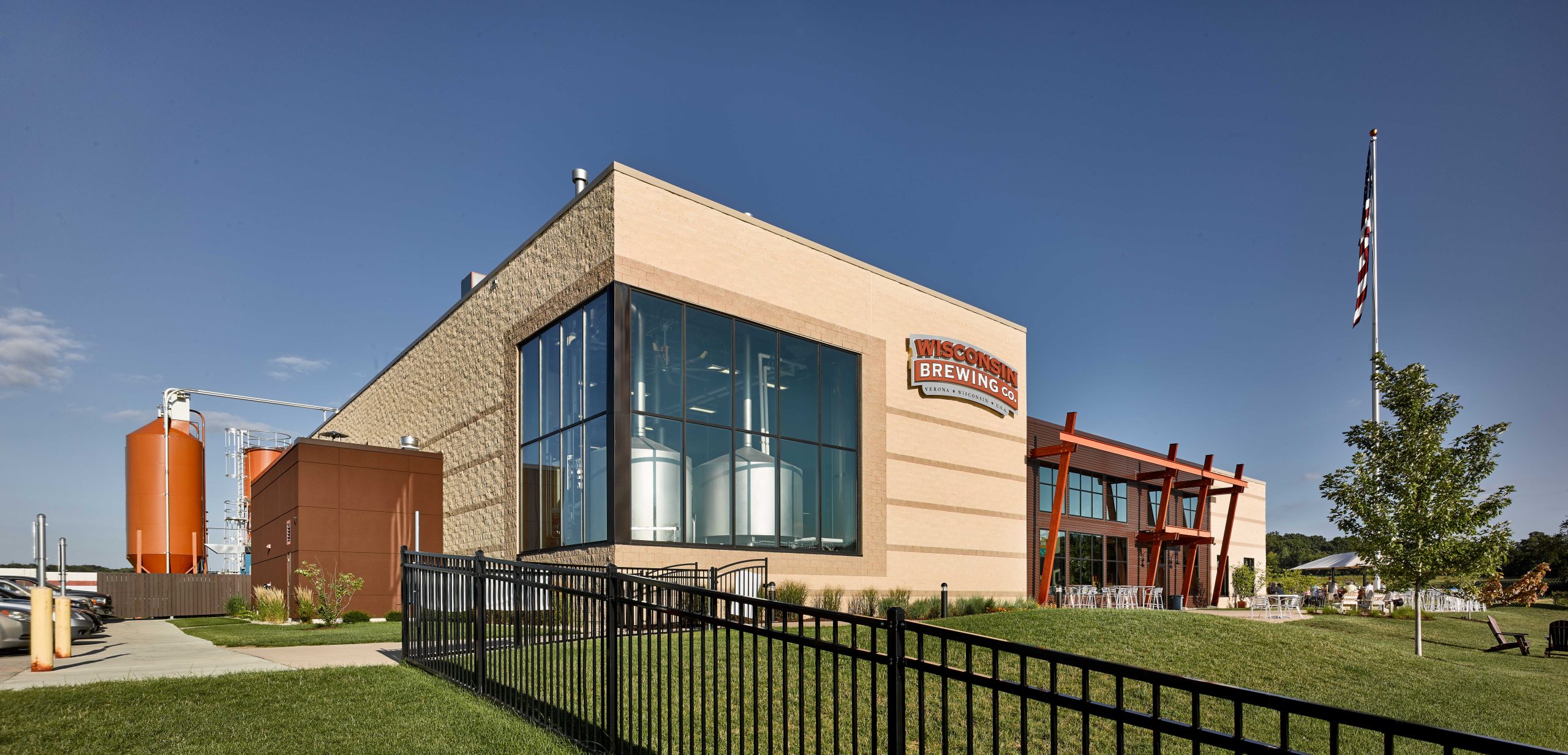 A modern industrial building with large windows and beige brick exterior stands near a pond with greenery. Several cars are parked in a lot beside the building. The sky is partly cloudy, creating soft, diffused light over the scene.