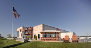 A large, modern building with a light exterior sits under a clear blue sky. An outdoor seating area with tables, chairs, and two large wooden armchairs is visible. A tall, prominent flagpole with an American flag waves in the foreground on a well-kept lawn.