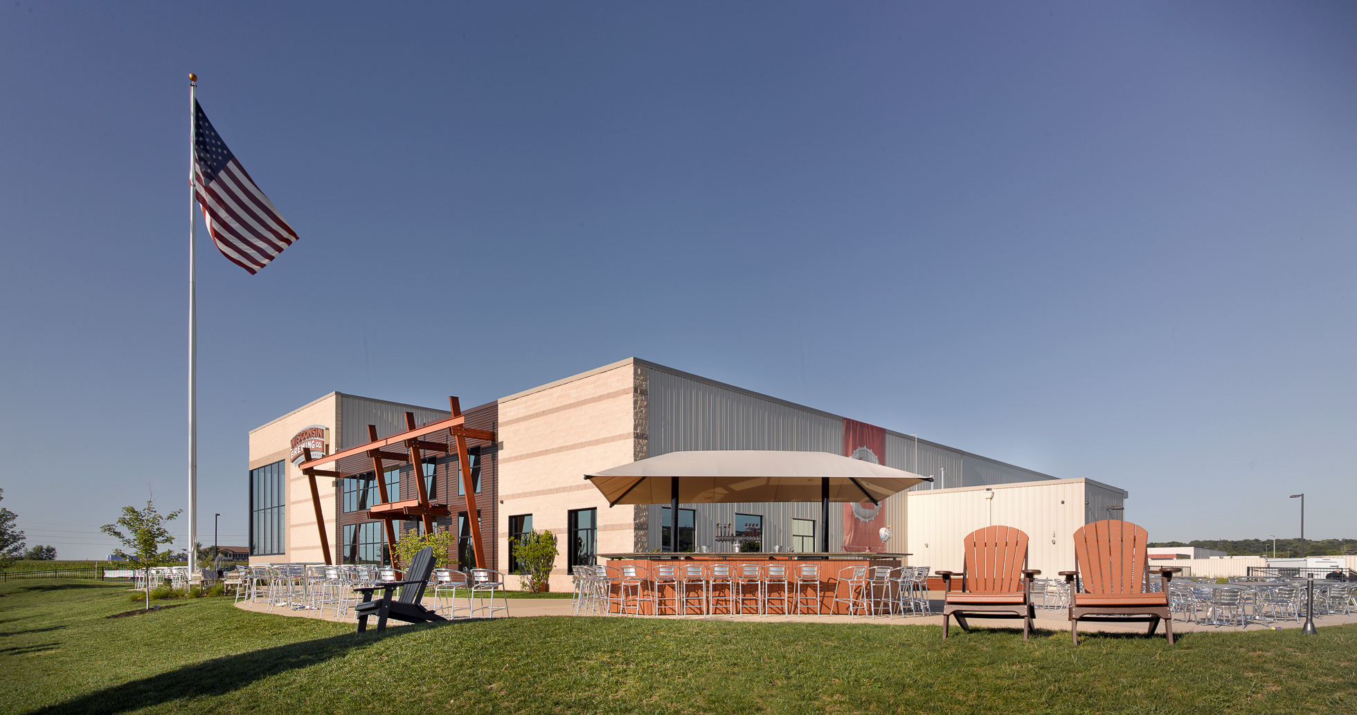 A large, modern building with a light exterior sits under a clear blue sky. An outdoor seating area with tables, chairs, and two large wooden armchairs is visible. A tall, prominent flagpole with an American flag waves in the foreground on a well-kept lawn.