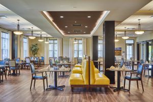 A spacious, brightly lit dining area with wooden floors and large windows. Yellow cushioned booths and neatly set tables are evenly spaced. The decor is modern with pendant lights and potted plants, creating a welcoming and orderly atmosphere.