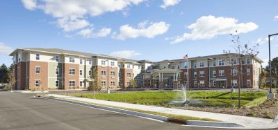 senior living facility exterior wide shot of the green, tan and red brick building with a pond and water fountain in front.