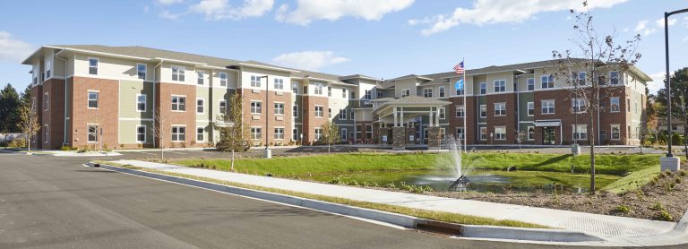 senior living facility exterior wide shot of the green, tan and red brick building with a pond and water fountain in front.