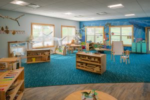 A spacious classroom with vibrant blue carpet featuring various learning stations and educational materials. Wooden furniture is scattered around, including shelves with toys and supplies, a small table with chairs, and an easel. Colorful decorations and artwork adorn the walls.