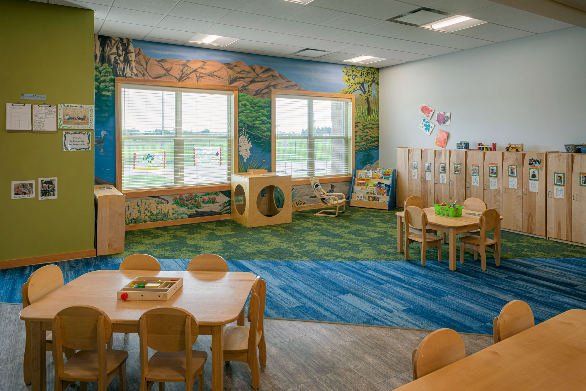 A brightly lit preschool classroom with windows, wooden tables, small chairs, and wall cubbies. The walls feature nature-themed murals with trees and mountains. The floor has blue and green carpet sections. Various toys and books are neatly arranged around the room.