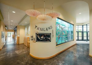 A brightly lit school hallway features a bulletin board and a wall display labeled "Thailand" with decorations, including a map and three hanging parasols. On the right is a large glass exhibit. The floor is polished and the ceiling has recessed lights.