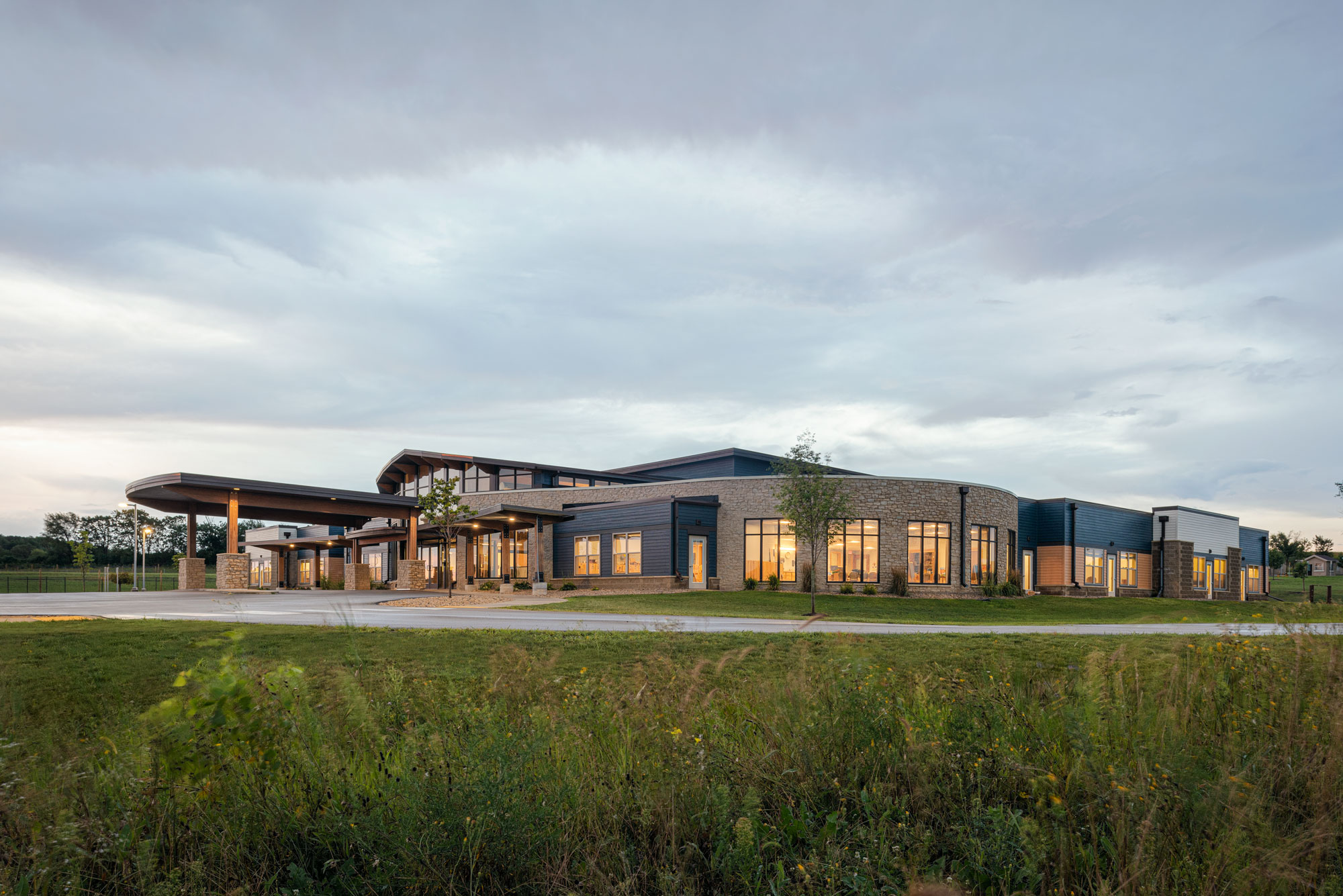 Modern, single-story building with large windows and a covered entryway, set against a cloudy sky at dusk. The structure features a mix of stone and metal exterior materials, and is surrounded by a grassy area with some small plants and trees.