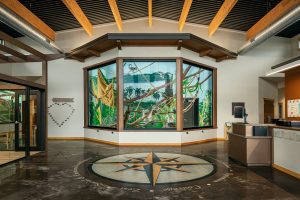 An indoor space with a polished floor featuring a large compass design in the center, wooden beams on the ceiling, and a colorful mural of tropical plants and landscape on the far wall. There's a heart-shaped design on the left wall and a reception desk to the right.