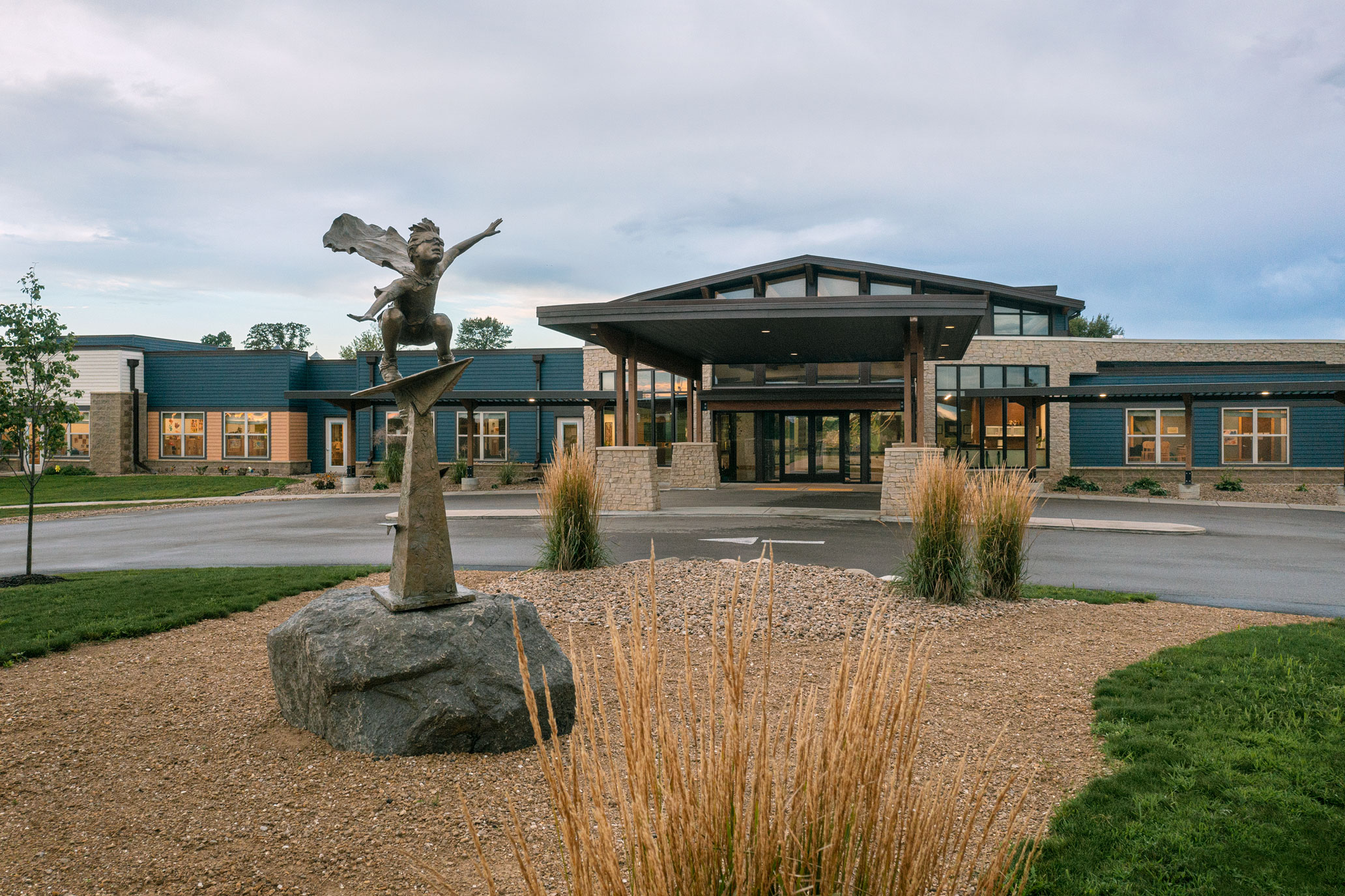 A modern building with a large entrance and a covered drop-off area. In the foreground, there is a stone sculpture of a person with outstretched arms standing on a rock, surrounded by tall grass and a landscaped area.